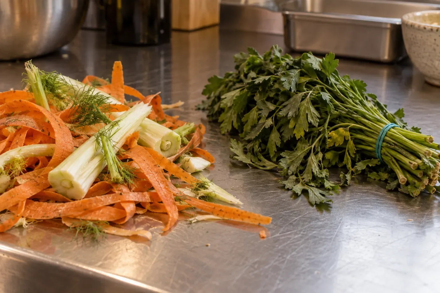 Banco di preparazione in acciaio con bucce di carota e gambi di finocchio appena pelati da un lato e un mazzo di prezzemolo legato con l'elastico dall'altro.